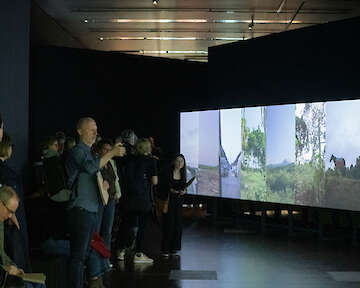 Ausstellungsansicht. Besucher*innen stehen vor einer großen Wand, auf die mehrere hochformatige Videos nebeneinander projiziert werden.  