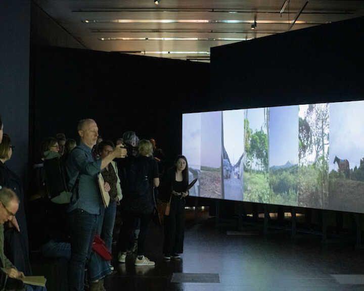 Ausstellungsansicht. Besucher*innen stehen vor einer großen Wand, auf die mehrere hochformatige Videos nebeneinander projiziert werden.  