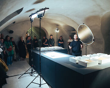 Pablo Lerma steht hinter einem Tisch vor Besuchenden und spricht in ein Mikro. Vor ihm stehen leere Boxen.  © buerofuerkunstdokumentation