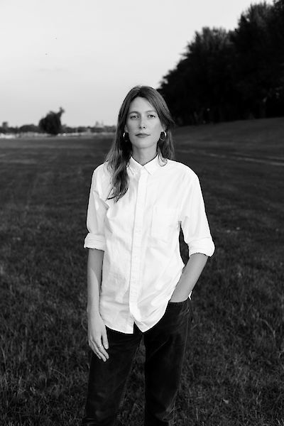 Portrait of Linda Conze in black and white. She is standing in a field, wearing long loose hair and a white shirt. One hand is in her trouser pocket.