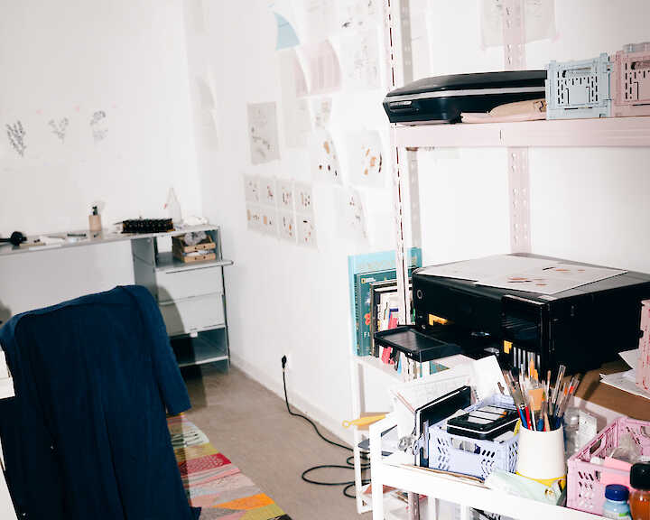 Blick in ein Atelier. Man sieht ein Regal mit verschiedenem Werkzeug und an der Wand hängen viele Bilder.  