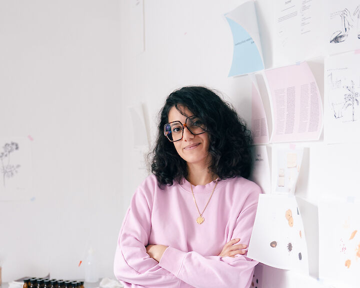 Portrait von Btihal Remli. Sie trägt eine Brille und einen rosa Pulli und lehnt an der Wand ihres Ateliers.  