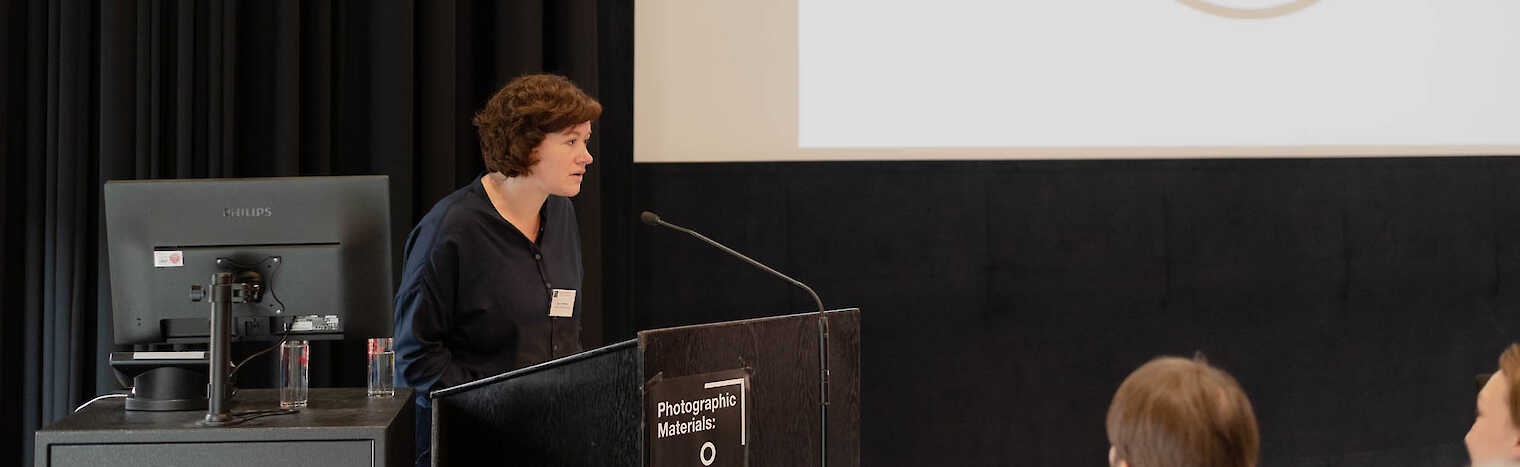 Inga Schneider steht an einem Sprechpult. Hinter ihr an einer Leinwand sieht man das Logo von Artist Meets Archive.