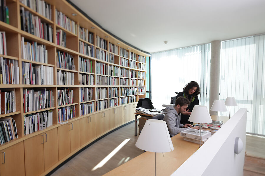 Zwei Personen beugen sich in der Bibliothek über ein Buch. Man sieht ein langes Regal voller Bücher und Arbeitsplätze.