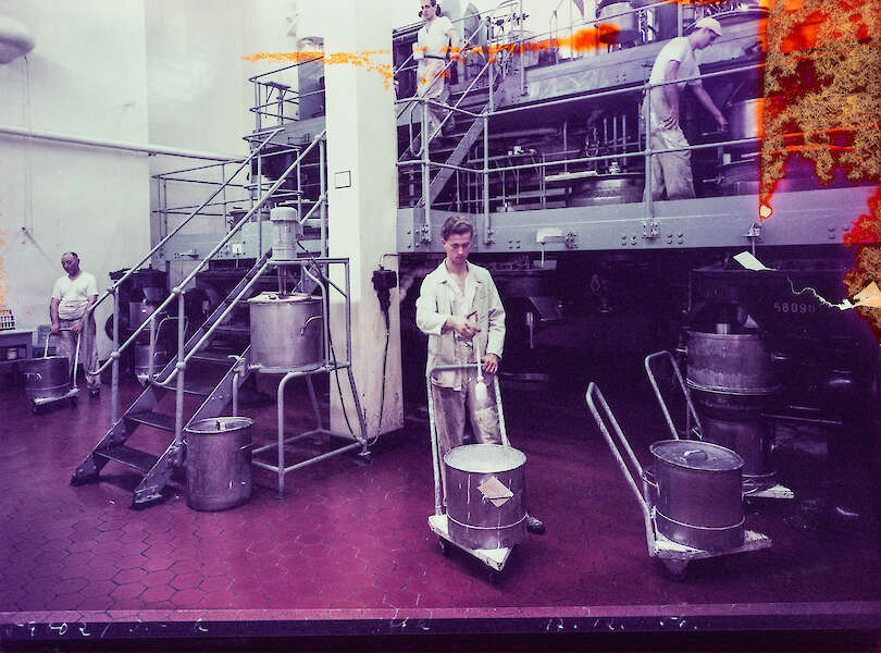 Arbeiter in einer Papierfabrik, die Maschinen bedienen und mit großen Fässern hantieren. Die Szene zeigt Metallgeräte und Treppen auf einem gefliesten Boden, der von einem violetten Farbton überlagert wird.