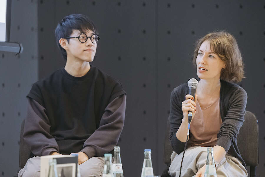 Jimmi Wing Kha Ho und Helena Weber sprechen in ein Mikrofon vor einer Leinwand