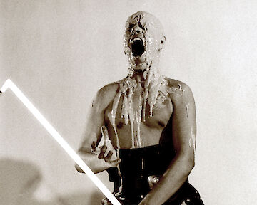 Eine Person sitzt auf einem Hocker, sie schreit und es sieht so aus, als wäre heißer Wachs auf das Gesicht und den Oberkörper getropft. Um die Beine sind Kabel von Stablampen gewunden. Eine Stablampe hält die Person vor sich in einer kämpferischen Haltung. Von Sinnen © Peter Rakoczy