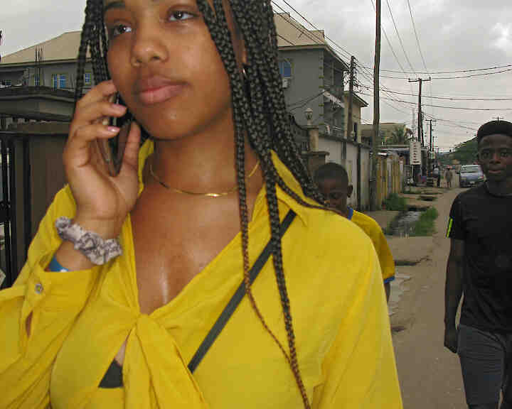 Eine schwarze Frau telefoniert mit ihrem Smartphone . Hnter ihr gehen zwei männlich gelesenen Personen. Einer davon ist noch jugenlich. Ada in Surulere © Anne Nwanneka