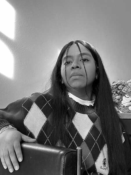 Portrait of G. Kühnhardt Alvarez in black and white. G. is wearing long loose hair and a sweater. Their hand is resting on the back of a chair.