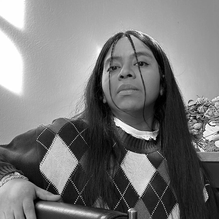 Portrait von G. Kühnhardt Alvarez in schwarz-weiß. G. trägt offenes langes Haar und einen Pullover. Deren Hand liegt auf einer Stuhllehne.  © Eva Scholl