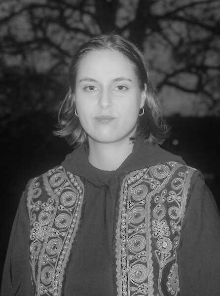 Portrait of Donja Nasseri in black and white. A dark tree can be seen in the background, Donja is flashed and looks into the camera. She is wearing a vest and a sweater.