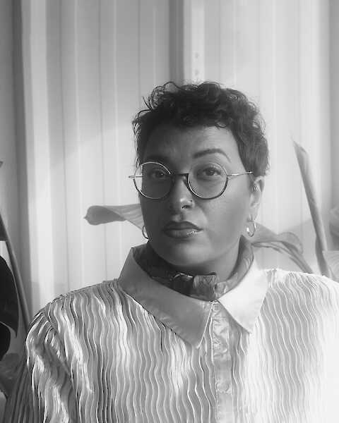 Portrait von Rebecca Racine Ramershoven in schwarz-weiß. Sie trägt eine runde Brille, kurze lockige Haare und eine weiße Bluse. Sie schaut in die Kamera.