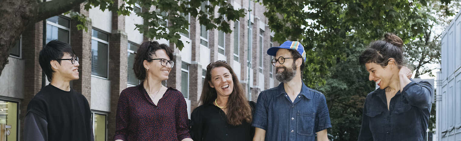 Die 5 Artist Meets Artist Künstler:innen stehen lächelnd auf einer gepflasterten Straße unter grünen Bäumen. Elena Efeoglou hat einen Dackel an der Leine.