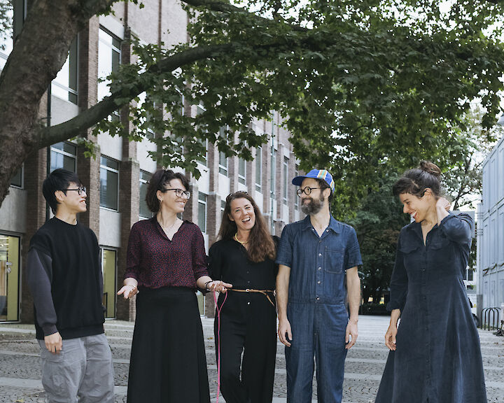 Die 5 Artist Meets Artist Künstler:innen stehen lächelnd auf einer gepflasterten Straße unter grünen Bäumen. Elena Efeoglou hat einen Dackel an der Leine.  © Silviu Guiman