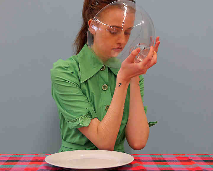 [Porträt einer weiblich gelesenen Person in apfelgrünem Kleid und spitzem Kragen im 70er Jahre Stil. Sie sitzt an einem Tisch mit karierter Tischdecke in verschiedenen Rottönen. Ihr Blick ist gesenkt und richtet sich in eine Glashaube, die sie mit ihren Händen vor ihr Gesicht hält. Die Person lächelt nicht. Ein leerer Teller steht vor der Person auf dem Tisch.] Friederike © Claudia Grünig