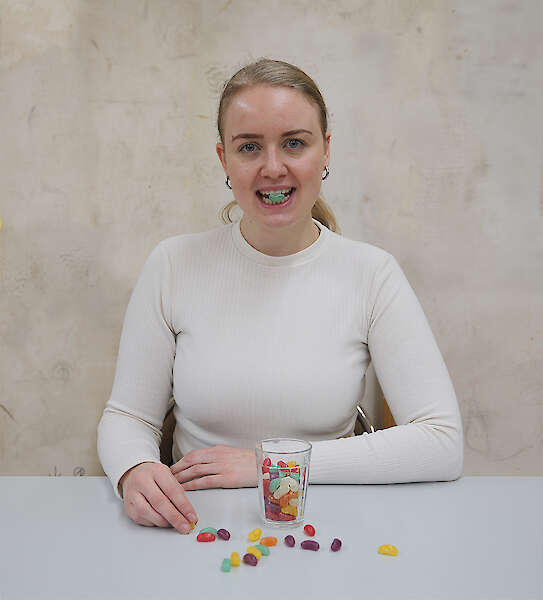 [Porträt einer weiblich gelesenen Person in weißem Oberteil. Die Person sitzt vor einem Glas buntem Fruchtgummi. Einige Fruchtgummi liegen auf der hellgrauen Tischplatte. Ein grünes Fruchtgummi steckt zwischen den Zahnreihen der Person. Sie schaut in die Kamera dabei.]