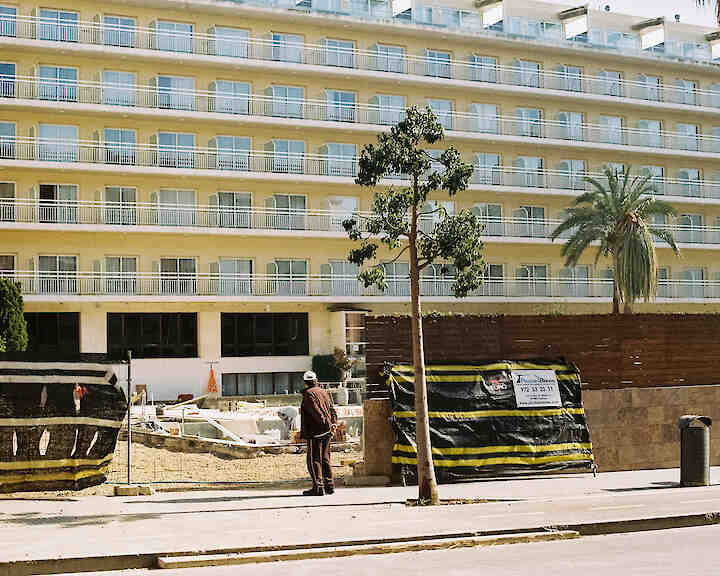 [Eine Person bleibt vor einem sehr großen Hotel in dem Urlaubsort Lloret de Mar, Mallorca, stehen. Die Hotelfassade füllt das gesamte Bild aus und zeigt sechs Stockwerke. Sichtbar sind zwanzig aneinander gereihte Fenster und Balkone. Das Hotel ist geschlossen. Ein Bauzaun grenzt das Grundstück ab. An dem Zaun hängt ein Schild mit der Telefonnummer einer Firma.] Lloret de Mar ©Caroline Brünen