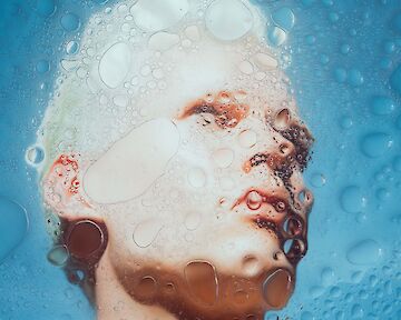 Ein Porträt einer Person hinter einer Fläche von Wassertropfen und Schaum. Es steht für unsere verzerrte Wahrnehmung von virtuellem Wasser. Virtuelles Wasser meint die Wassermengen, die entlang der gesamten Wertschöpfungskette von Produkten benötigt wird.  © Jule Petersen