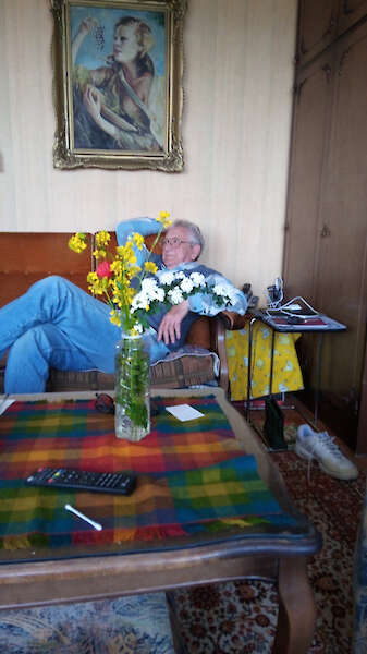 Ein älterer Mann entspannt sich auf einer Couch in einem Wohnzimmer und stützt sich auf ein Kissen. Eine Vase mit gelben und weißen Blumen steht auf einem Tisch mit einem bunten Tuch. An der Wand hinter ihm hängt ein Gemälde einer Frau, und neben einem Beistelltisch stehen Schuhe.