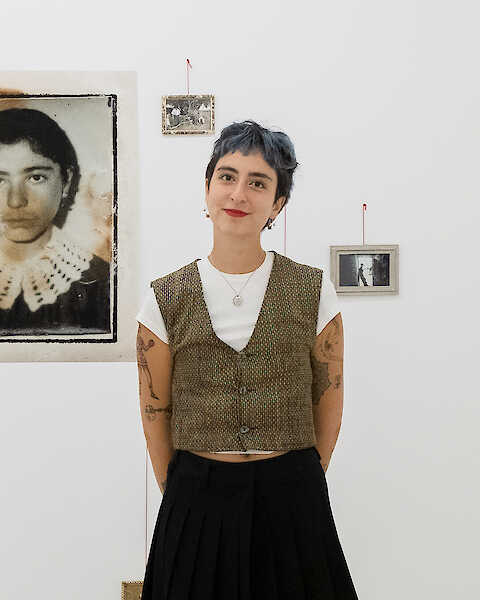 Die Künstlerin steht in ihrer Ausstellung. Sie trägt roten Lippenstift und eine Weste.