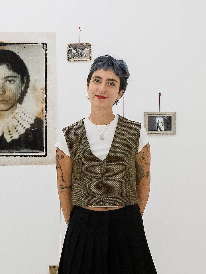 Die Künstlerin steht in ihrer Ausstellung. Sie trägt roten Lippenstift und eine Weste.  © Aslı Özçelik