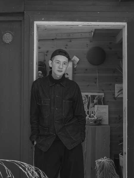 Künstlerportrait: Ein dunkel gekleideter Mann mit Mütze und Brille lehnt an einem Türrahmen. Der Raum dahinter ist holzgetäfelt und mit verschiedenen Gegenständen und Kunstwerken in Regalen ausgestattet. Das Bild ist schwarz-weiß.