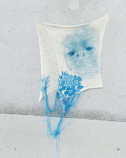 Ein durchscheinendes Stoffkunstwerk mit einem blassen, abstrakten Gesicht und blauen Blumenmustern ist an einer Betonwand angebracht. Blaue Schnüre baumeln vom Stoff herab.