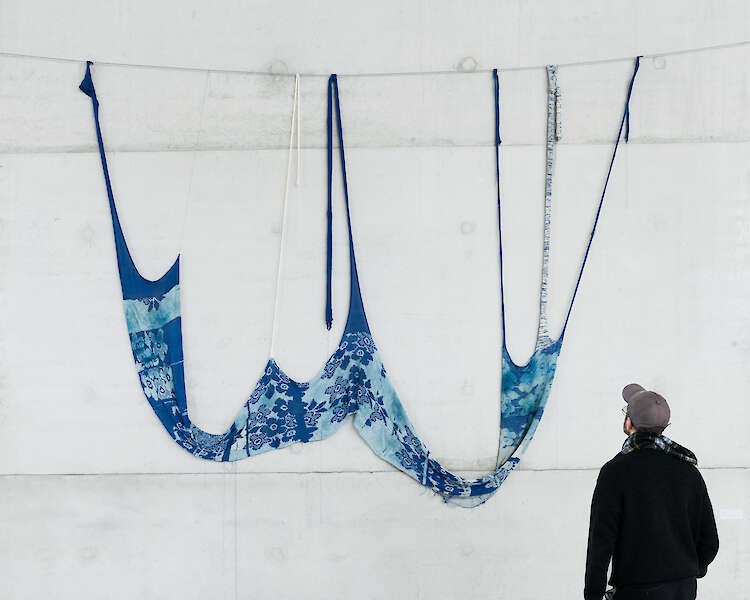 Eine Person blickt auf eine Kunstinstallation mit langen blauen Stoffstreifen, die an einer weißen Wand hängen. Der Stoff weist verschiedene Muster und Blautöne auf und bildet eine wellenförmige, schwebende Form.