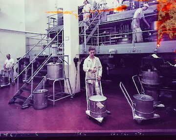 Workers in a paper factory operating machinery and handling large barrels. The scene includes metal equipment and staircases against a tiled floor, with a purple hue overlay. Fotograf:in unbekannt, Barytküche, 17.12.1956, Farbfotografie, Museum Ludwig, Agfa Werbearchiv © Fotograf*in unbekannt, Agfa Werbearchiv, Museum Ludwig
