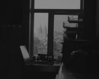 Schwarz-Weiße Innenaufnahme aus einem dunklen Raum, mit Blick auf die Skyline einer Stadt durch ein Fenster. "The View of St. Michaels Cathedral", Qingdao, 2024 © Jimmi Wing Ka Ho