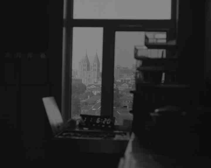 Schwarz-Weiße Innenaufnahme aus einem dunklen Raum, mit Blick auf die Skyline einer Stadt durch ein Fenster. "The View of St. Michaels Cathedral", Qingdao, 2024 © Jimmi Wing Ka Ho