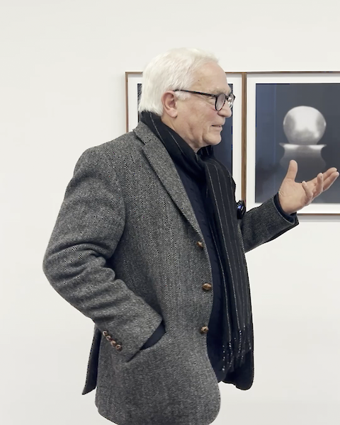 Norbert Moos steht vor einer Galeriewand mit eingerahmten Fotos. Er trägt ein graues Jacket und gestikuliert mit einer Hand.