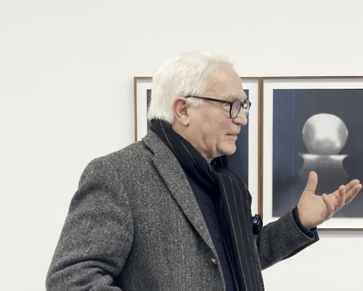 Norbert Moos steht vor einer Galeriewand mit eingerahmten Fotos. Er trägt ein graues Jacket und gestikuliert mit einer Hand.  