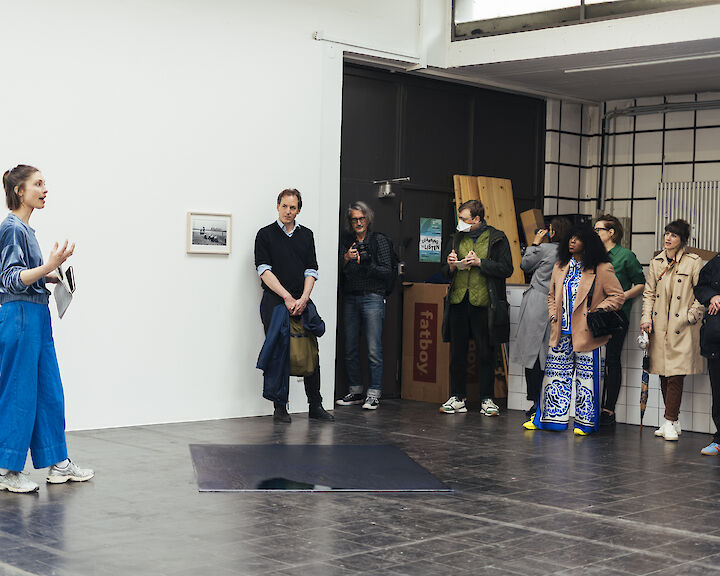Eine Person in blauer Hose steht vor einer Gruppe von Menschen in einem Ausstellungsraum. Sie erzählt ihnen etwas.  © buerofuerkunstdokumentation